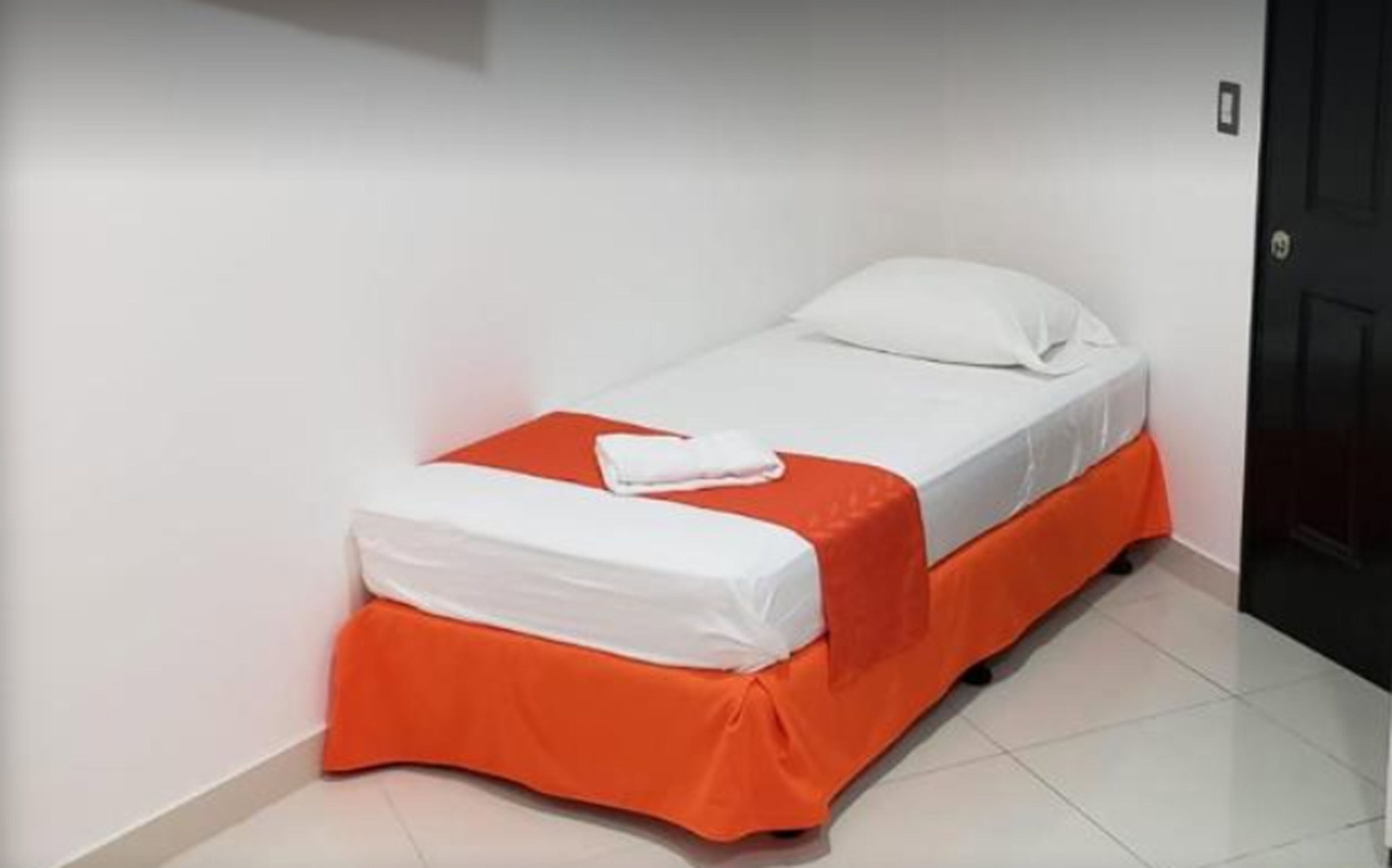 A small, single-bed room with white bedding and an orange skirt.
