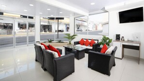 A modern hotel lobby with wicker furniture, a flat-screen TV, and a view of the street through large glass doors. - Hotel Milan Costa Rica (San José)