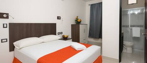 Habitación de hotel con cama, mesita de noche, armario y baño visible a través de una puerta abierta.