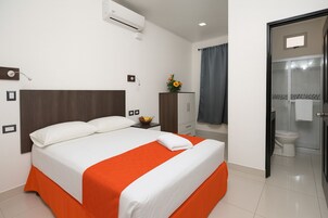 Habitación de hotel con cama, mesita de noche, armario y baño visible a través de una puerta abierta.
