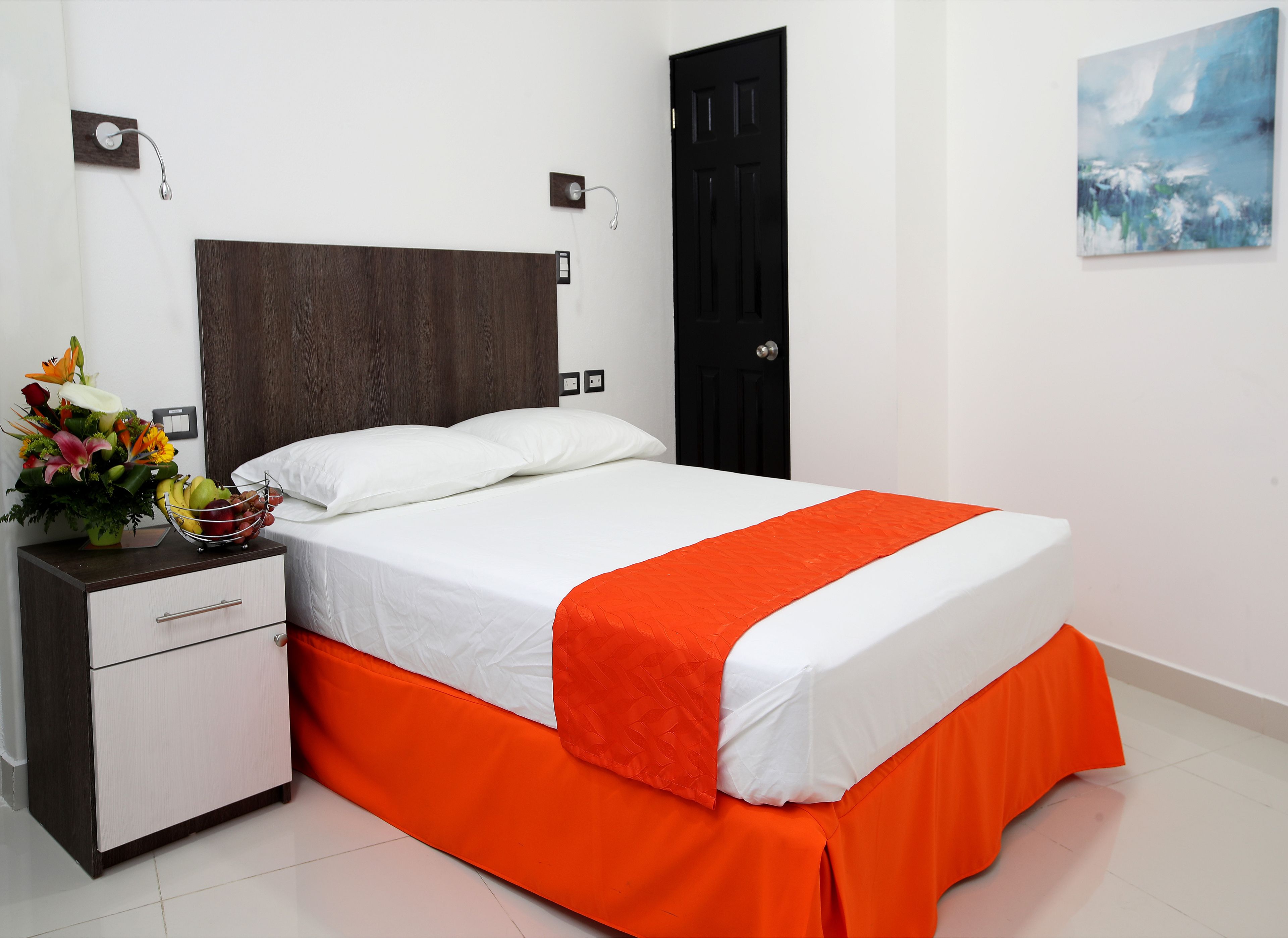Uma cama bem arrumada com lençóis brancos e saia laranja, uma mesa de cabeceira com um vaso de flores, uma porta preta e um quadro na parede.