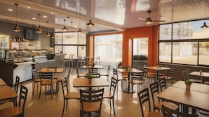 A modern cafe with wooden tables and chairs, a counter with a display case, and large windows allowing natural light. - Hotel Milan Costa Rica (San José)