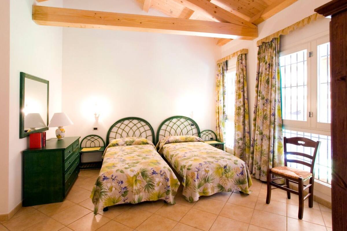 Ein Schlafzimmer mit zwei Einzelbetten, einer Holzbalkendecke, einer grünen Kommode, einem Stuhl und einem Fenster mit Vorhängen.