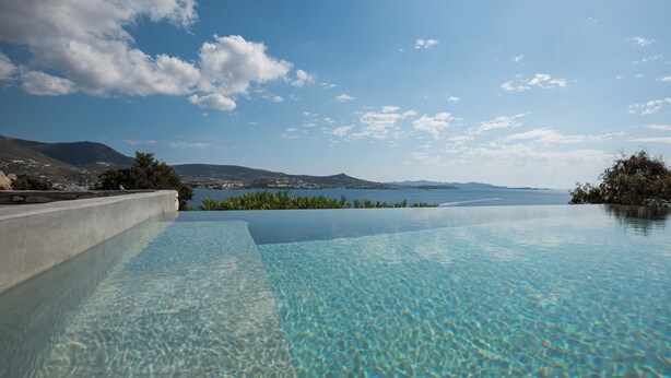 En infinitypool med kristallklart vatten, omgiven av en betongkant, med en vacker utsikt över en sjö och avlägsna berg under en delvis molnig himmel.