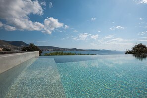 An infinity pool with clear water, surrounded by a concrete edge, overlooking a scenic view of a lake and distant mountains under a partly cloudy sky. - Krios Luxury Suites (Paros)