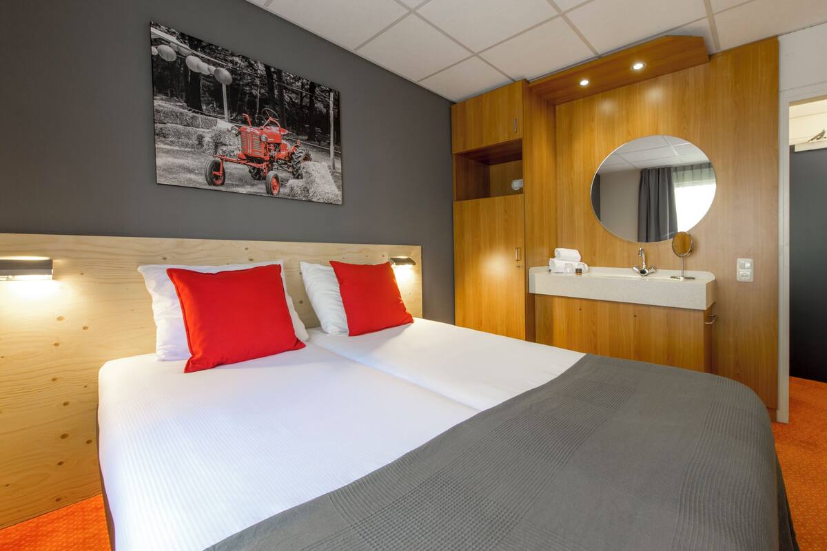 A hotel room with a large bed, a wooden headboard, a round mirror, and a wall-mounted picture of a tractor.