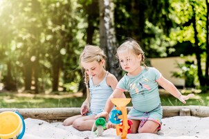 Dos niños jugando con arena y juguetes al aire libre, con árboles de fondo.