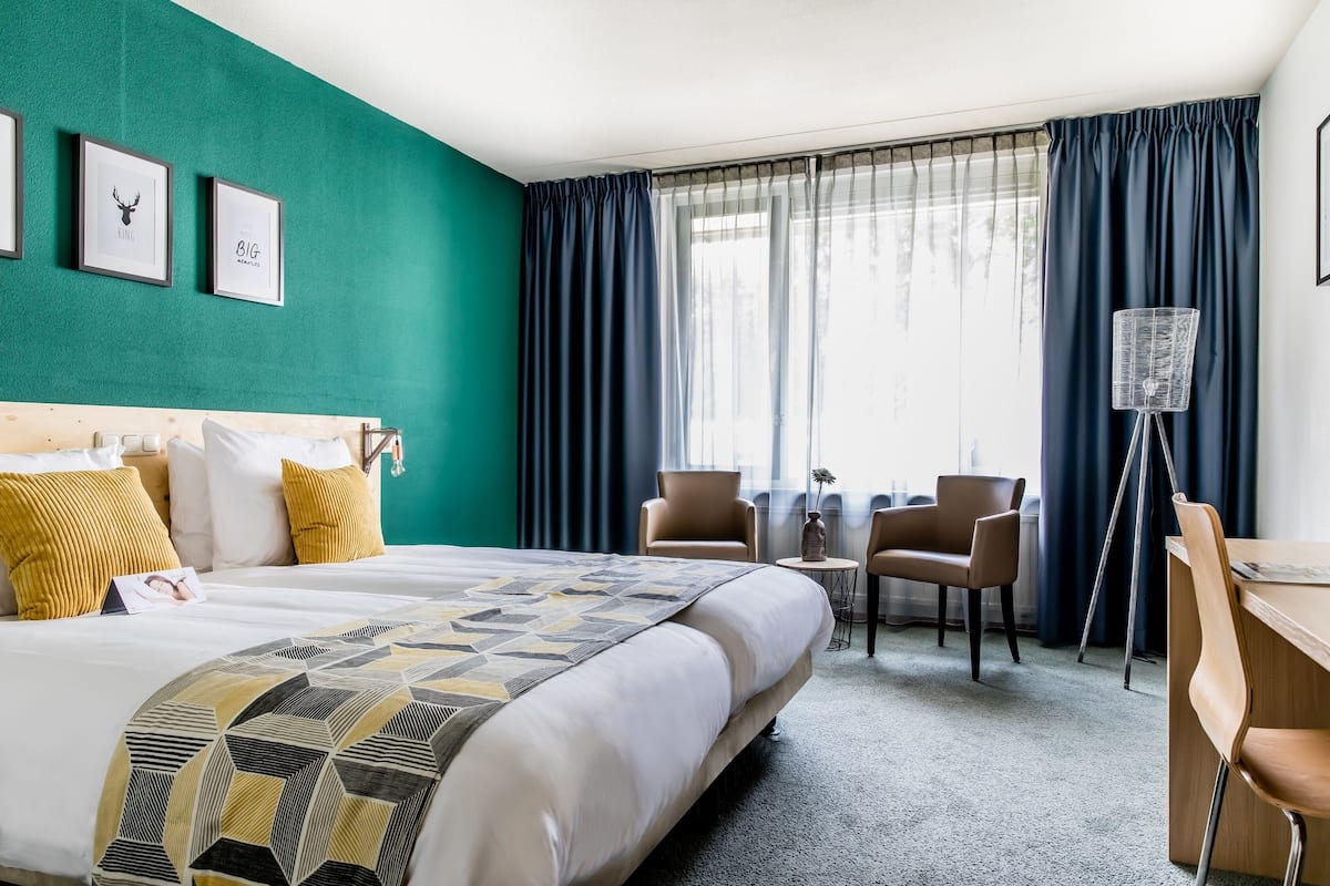 A hotel room with a large bed, a desk, two chairs, a small table, and a floor lamp.