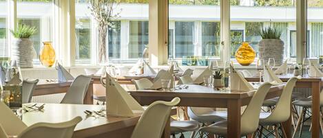 Un comedor con varias mesas y sillas, ventanas amplias con vistas a la vegetación y jarrones decorativos en los alféizares.