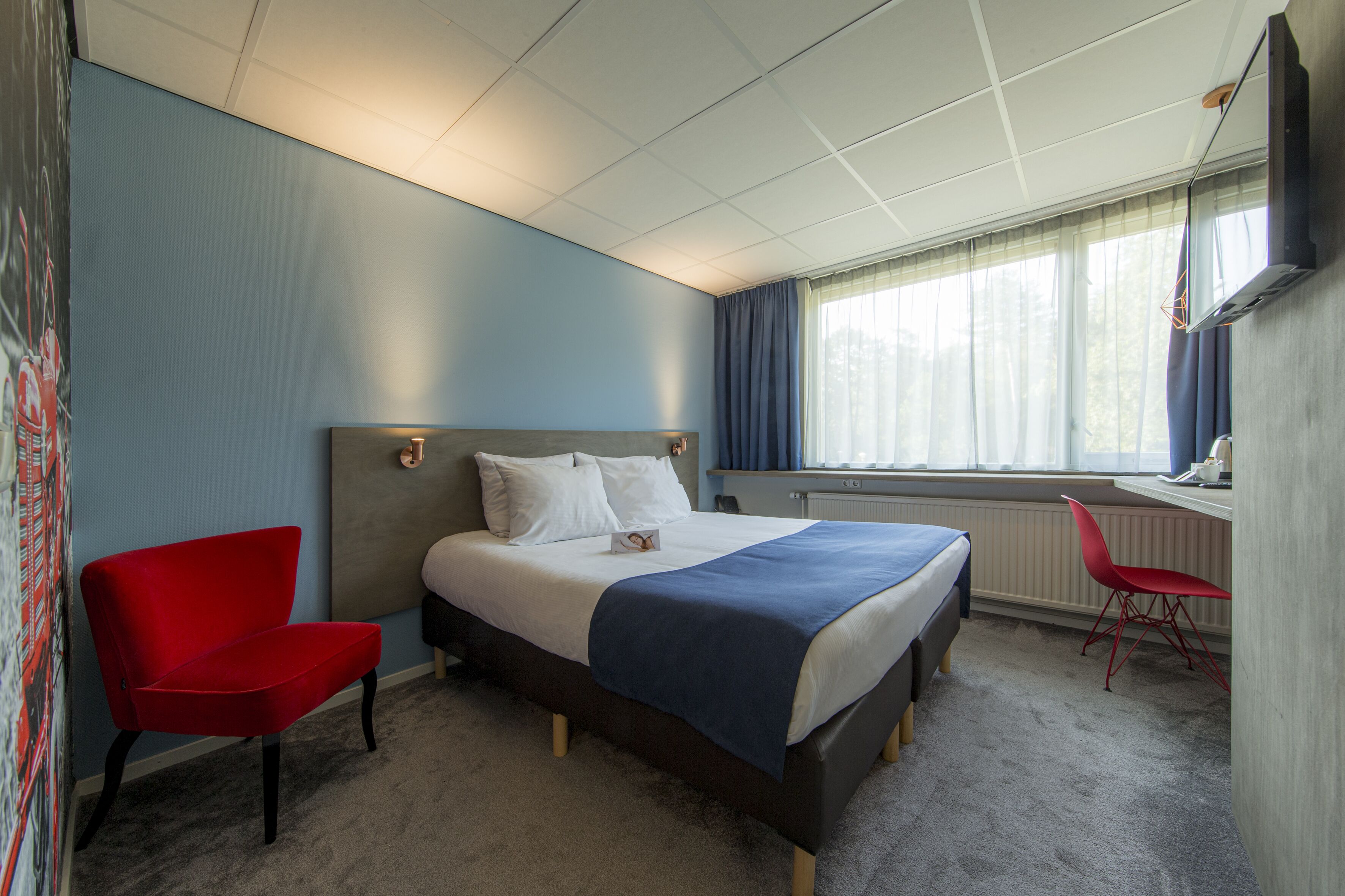 A hotel room with a bed, a red chair, a desk, and a window with curtains.