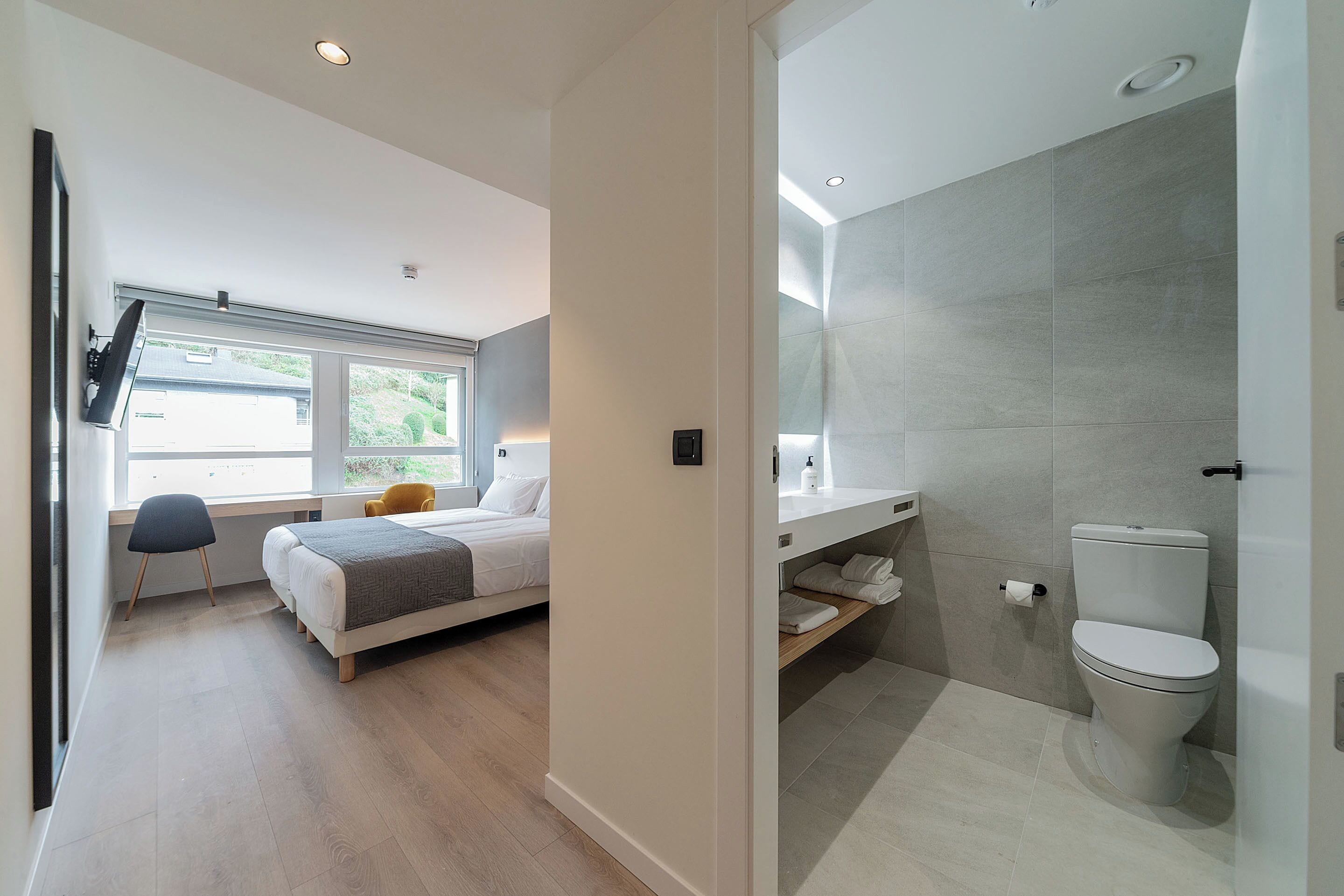 A modern hotel room with a bed, a desk, a chair, a window, and a bathroom with a toilet and sink.