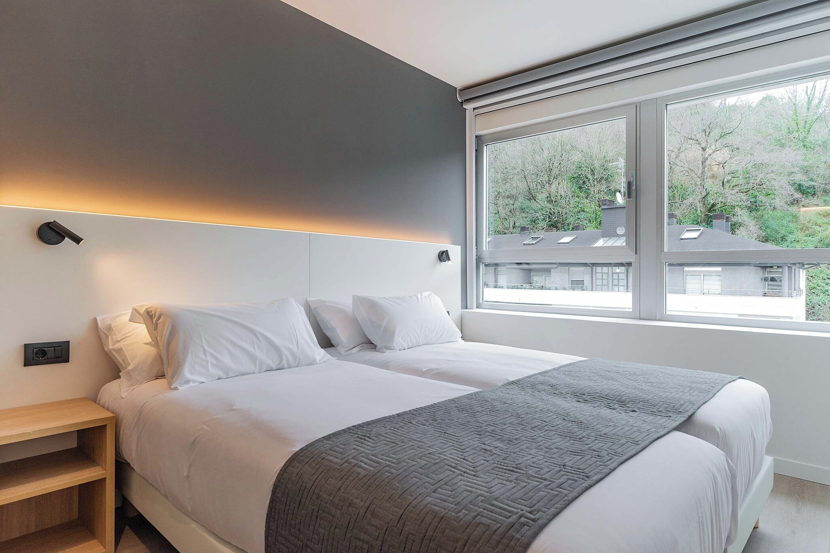 A modern bedroom with a bed, bedside tables, a window with a view of trees, and a wall-mounted light.