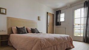 A bedroom with a wooden headboard, a bed with a pink bedspread, a nightstand, a window with curtains, and a door leading to another room. - Hôtel Galilee (Saint-Michel-l'Observatoire)