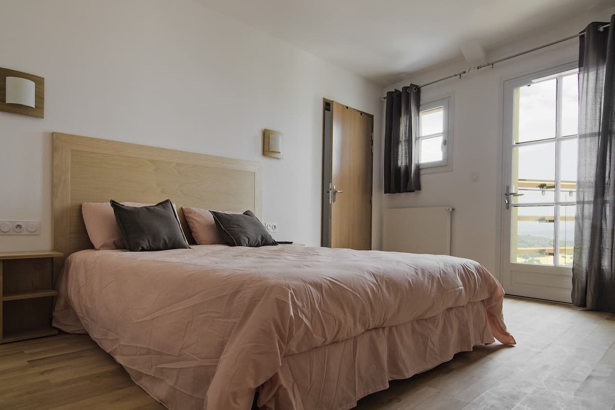 Een slaapkamer met een houten hoofdbord, een bed met een roze dekbedovertrek, een nachtkastje, een raam met gordijnen en een deur naar een andere kamer.