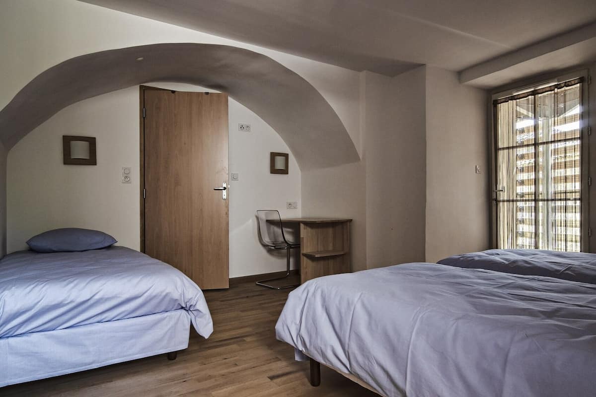 Een kamer met twee bedden, een houten deur, een klein tafeltje en een raam met jaloezieën.
