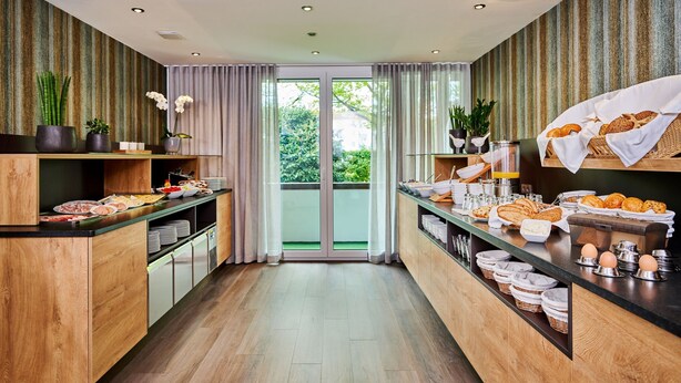 A modern breakfast bar with a variety of food items, including bread, pastries, and eggs, with a view of greenery outside. - City Appartementhotel (Bad Fuessing)