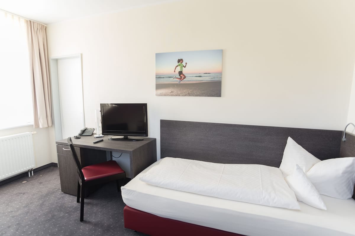 Ein Hotelzimmer mit einem Bett, einem Schreibtisch mit Computer, einem Stuhl und einem Fenster mit Vorhängen.