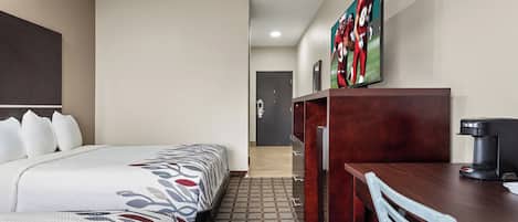 A hotel room with a bed, a desk with a coffee maker, a TV on the wall, and a door in the background.