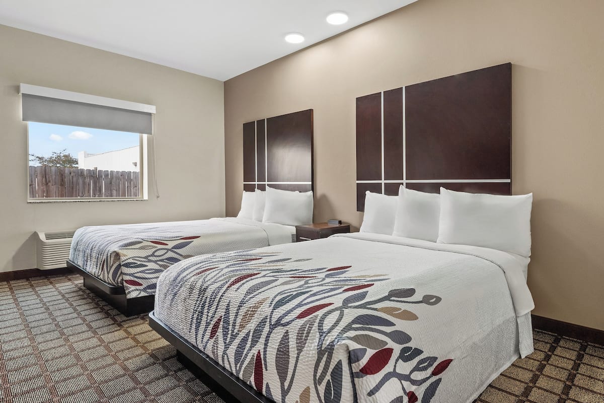 A hotel room with two beds, a large window with a view of a fence, and a patterned carpet.