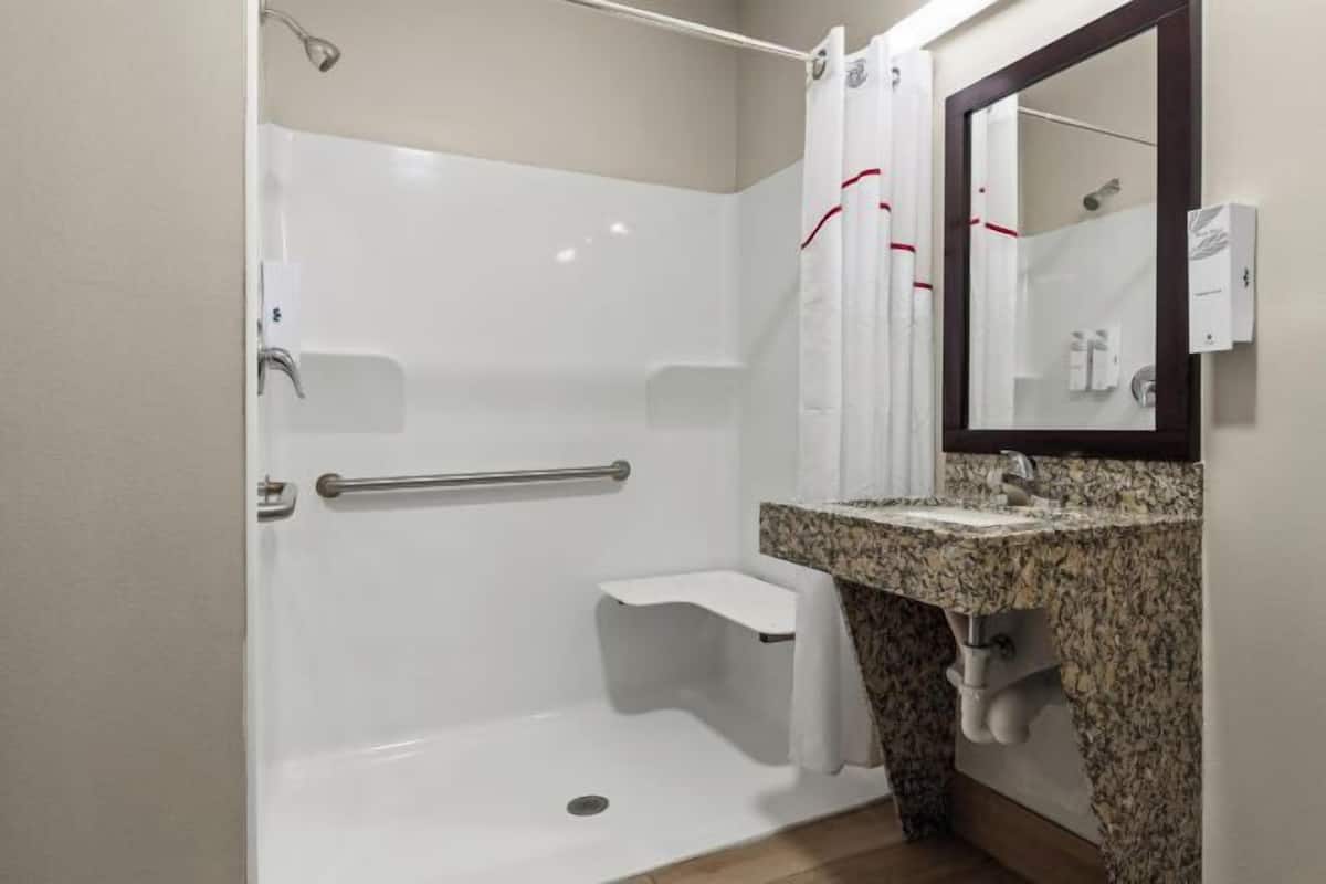 A bathroom with a walk-in shower, a grab bar, a sink with a granite countertop, and a mirror with a towel hanging on it.