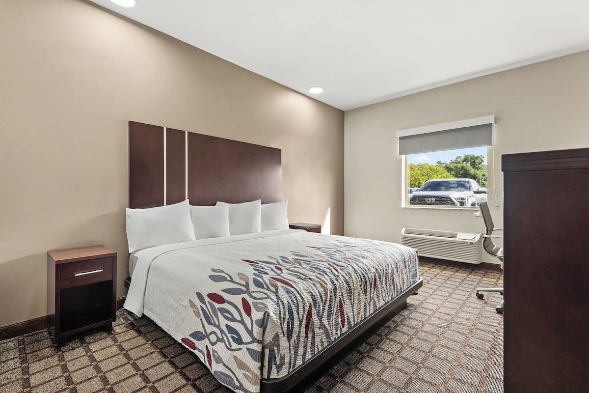 A hotel room with a large bed, a nightstand, a window with a view of a car, and an air conditioning unit.