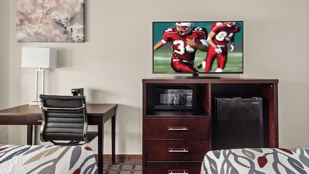 A hotel room with a TV displaying a football game, a desk with a chair, a bed with patterned bedding, and a microwave in a cabinet. - Red Roof Inn Baton Rouge (Baton Rouge)