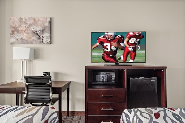 A hotel room with a TV displaying a football game, a desk with a chair, a bed with patterned bedding, and a microwave in a cabinet.