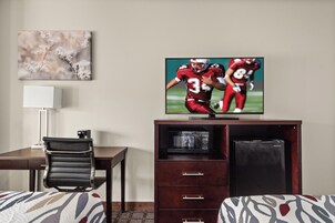 Habitación de hotel con una televisión mostrando un partido de fútbol, un escritorio con una silla, una cama con ropa de cama estampada y un microondas en un armario.