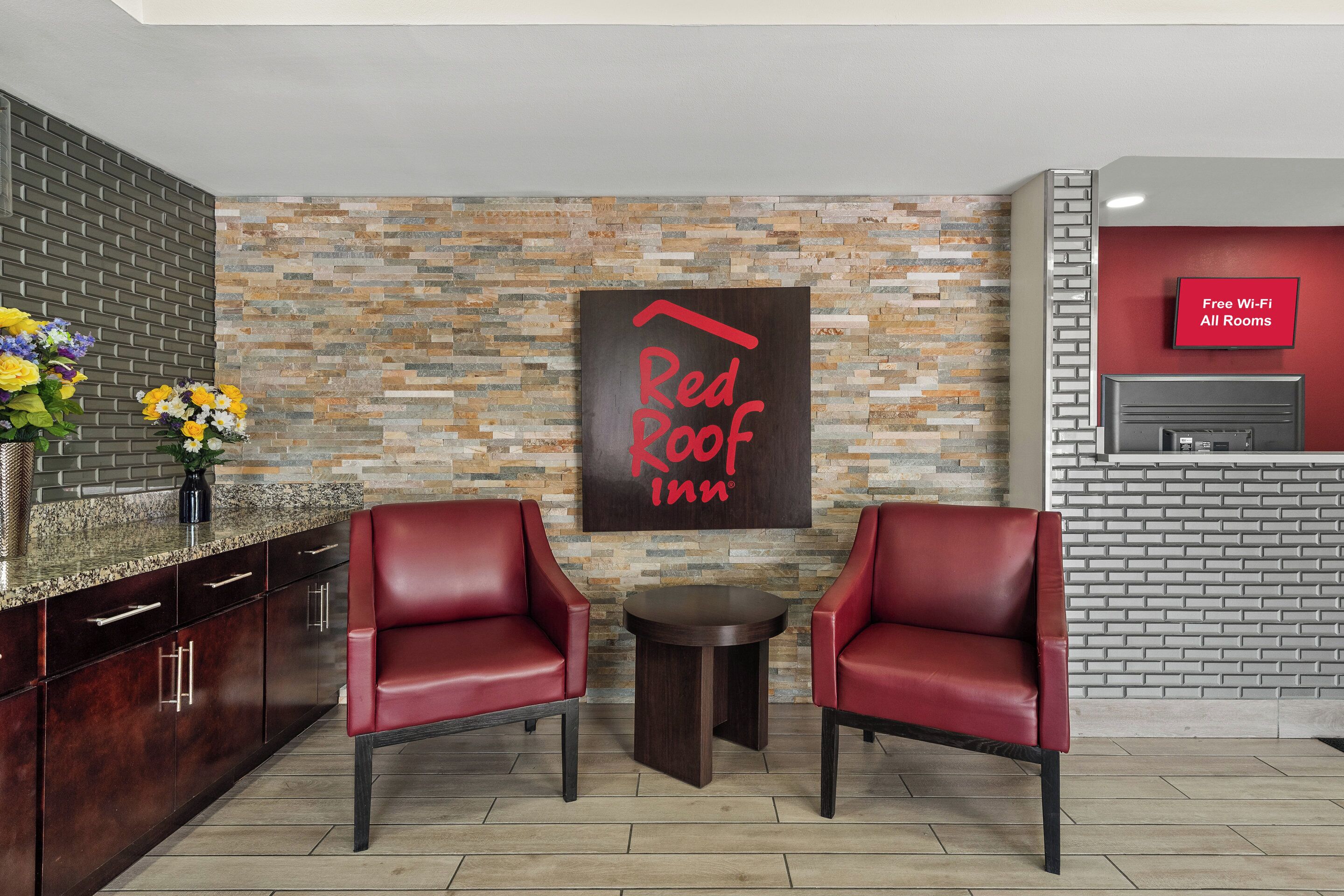 Die Lobby eines Red Roof Inn mit Backsteinwand, zwei roten Ledersesseln, einem kleinen runden Tisch und einem Fernseher, der „Kostenloses WLAN in allen Zimmern“ anzeigt.