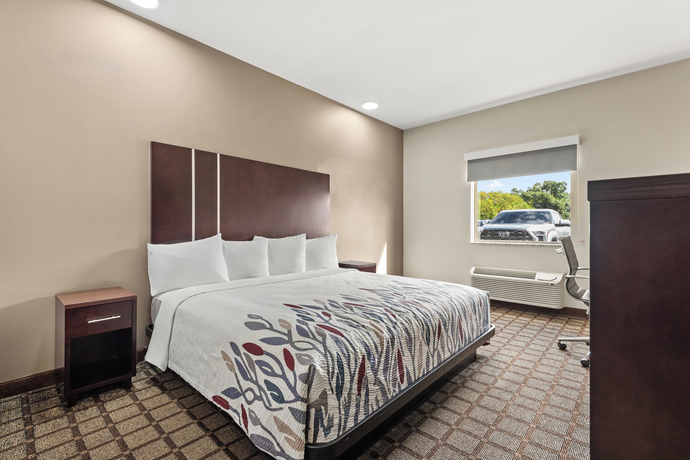 A hotel room with a large bed, a nightstand, a window with a view of a car, and an air conditioning unit.