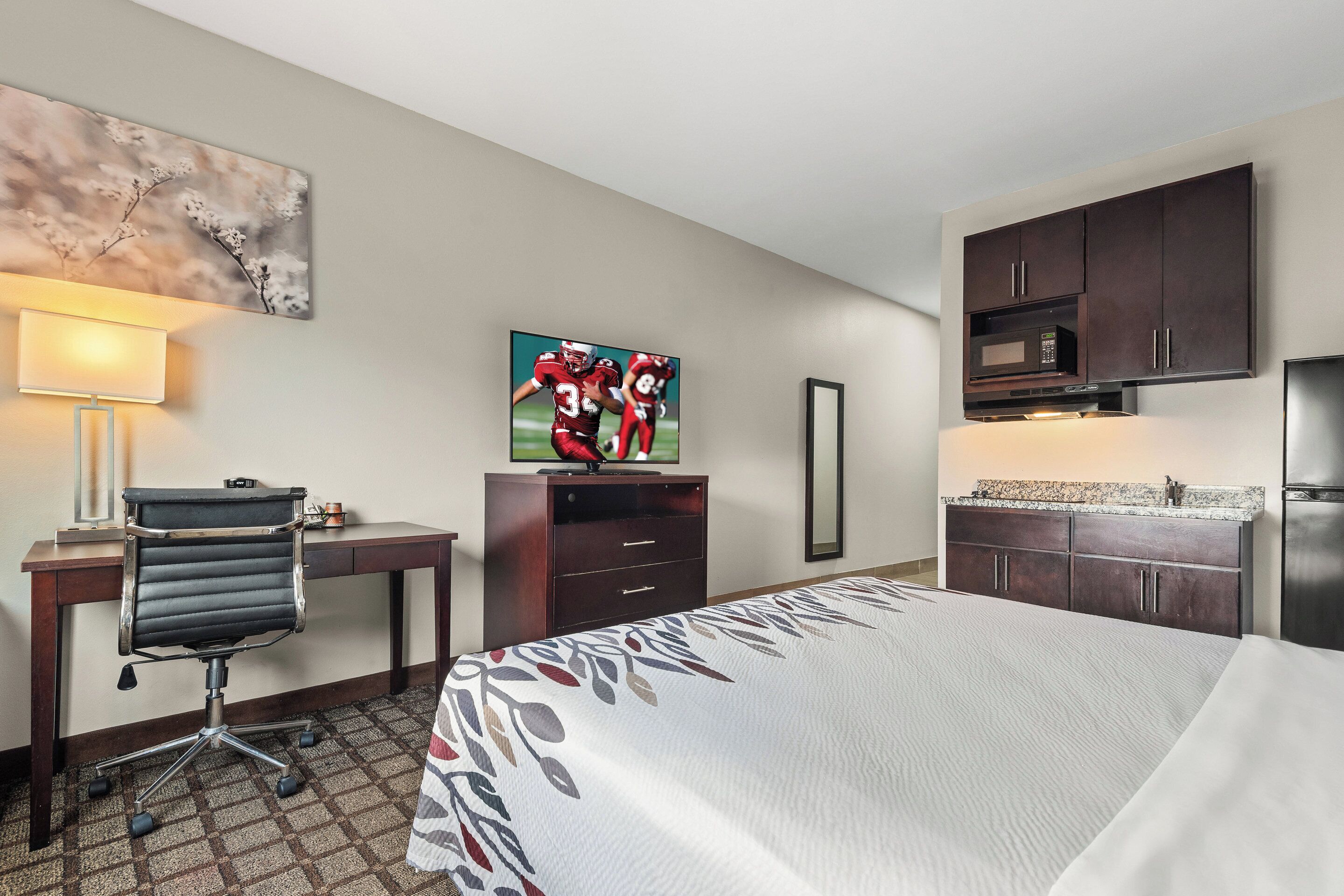 A hotel room with a bed, desk, chair, TV, microwave, and kitchenette.