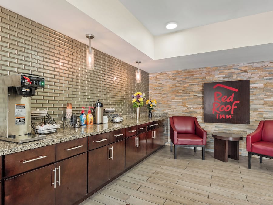 A modern kitchen with a coffee machine, a sink, and a countertop with various items. There are two red chairs and a small table. A sign on the wall reads "Red Roof Inn."