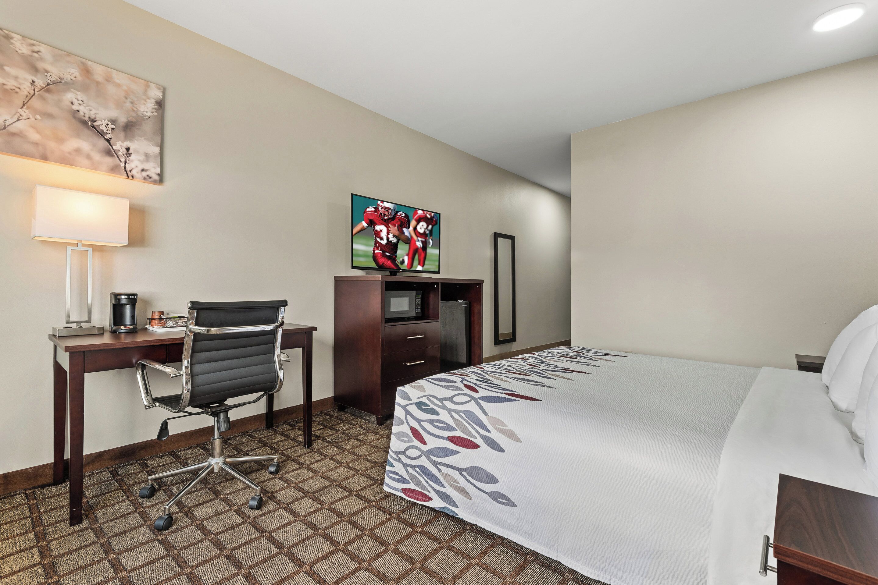 A hotel room with a bed, a desk with a chair, a TV, and a lamp.
