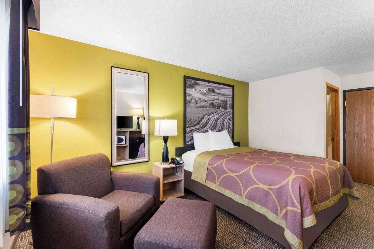 A hotel room with a bed, two armchairs, a nightstand with lamps, a mirror, and a wall-mounted picture.
