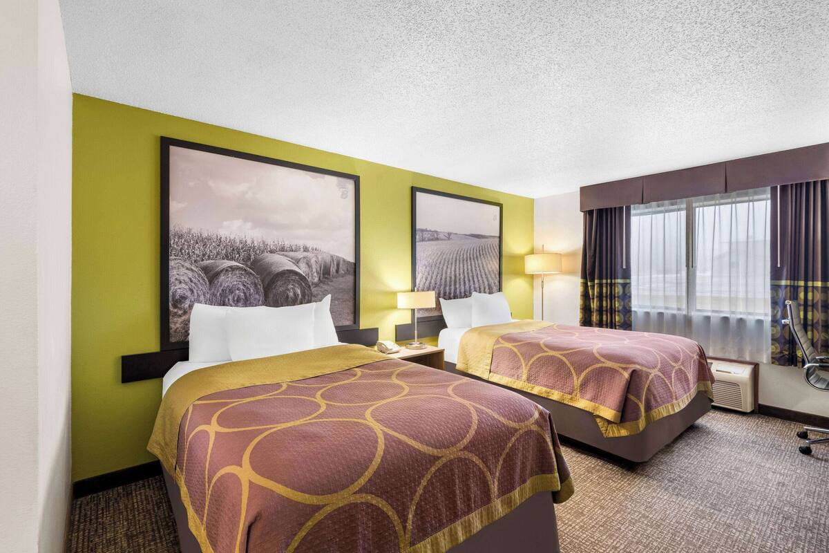 A hotel room with two beds, a large window with curtains, a lamp, and two framed pictures on the wall.