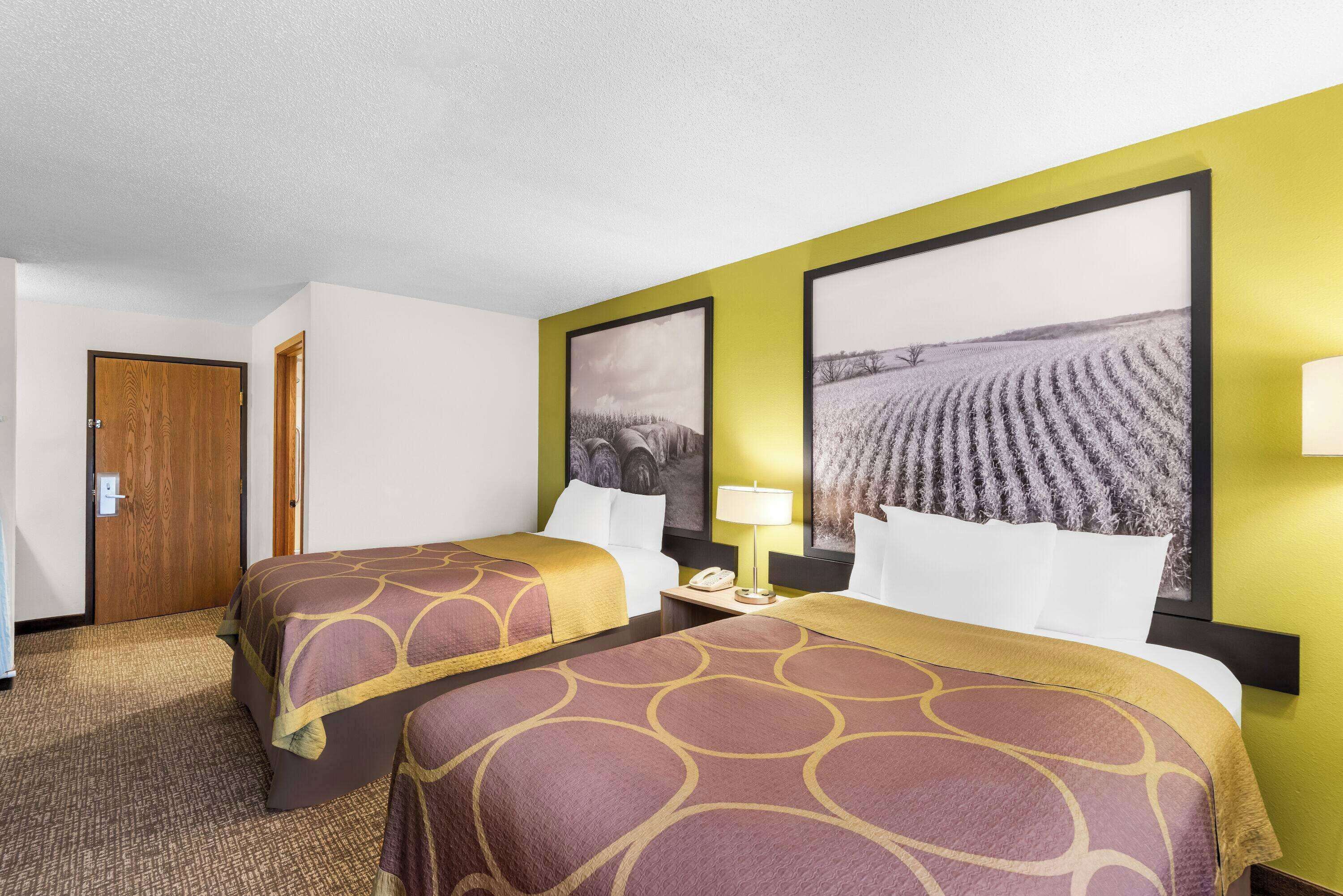 Quarto de hotel com duas camas, uma porta de madeira e duas grandes pinturas de paisagens nas paredes.