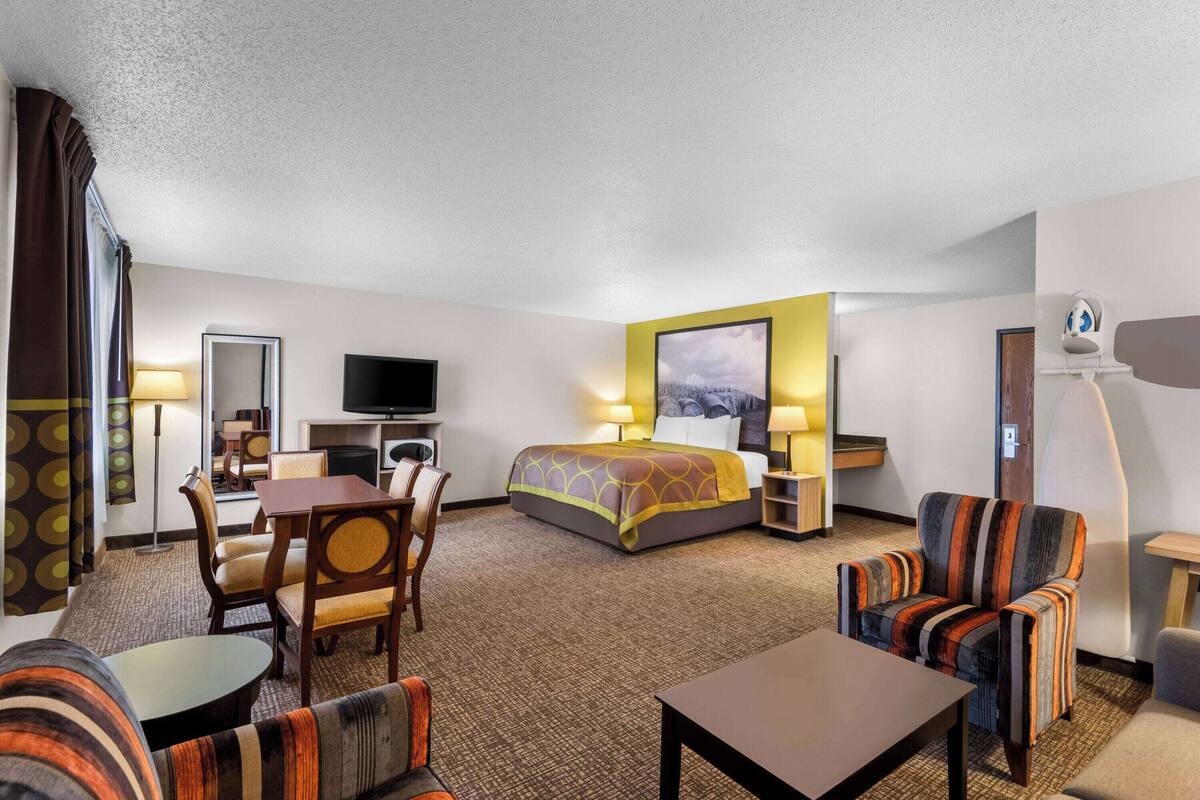 A hotel room with a bed, a desk, two chairs, a sofa, a TV, and a painting on the wall.