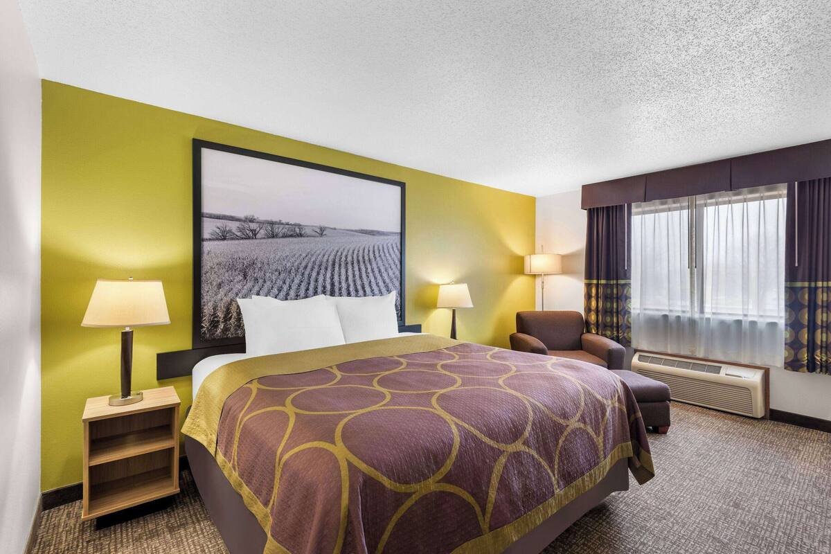 A hotel room with a large bed, two bedside lamps, a chair, a window with curtains, and a wall-mounted air conditioner.