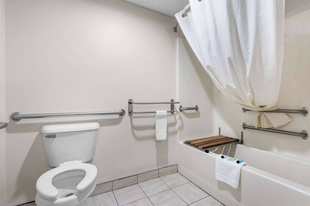 A bathroom with a white toilet, grab bars, a wooden seat, a bathtub, and a shower curtain.