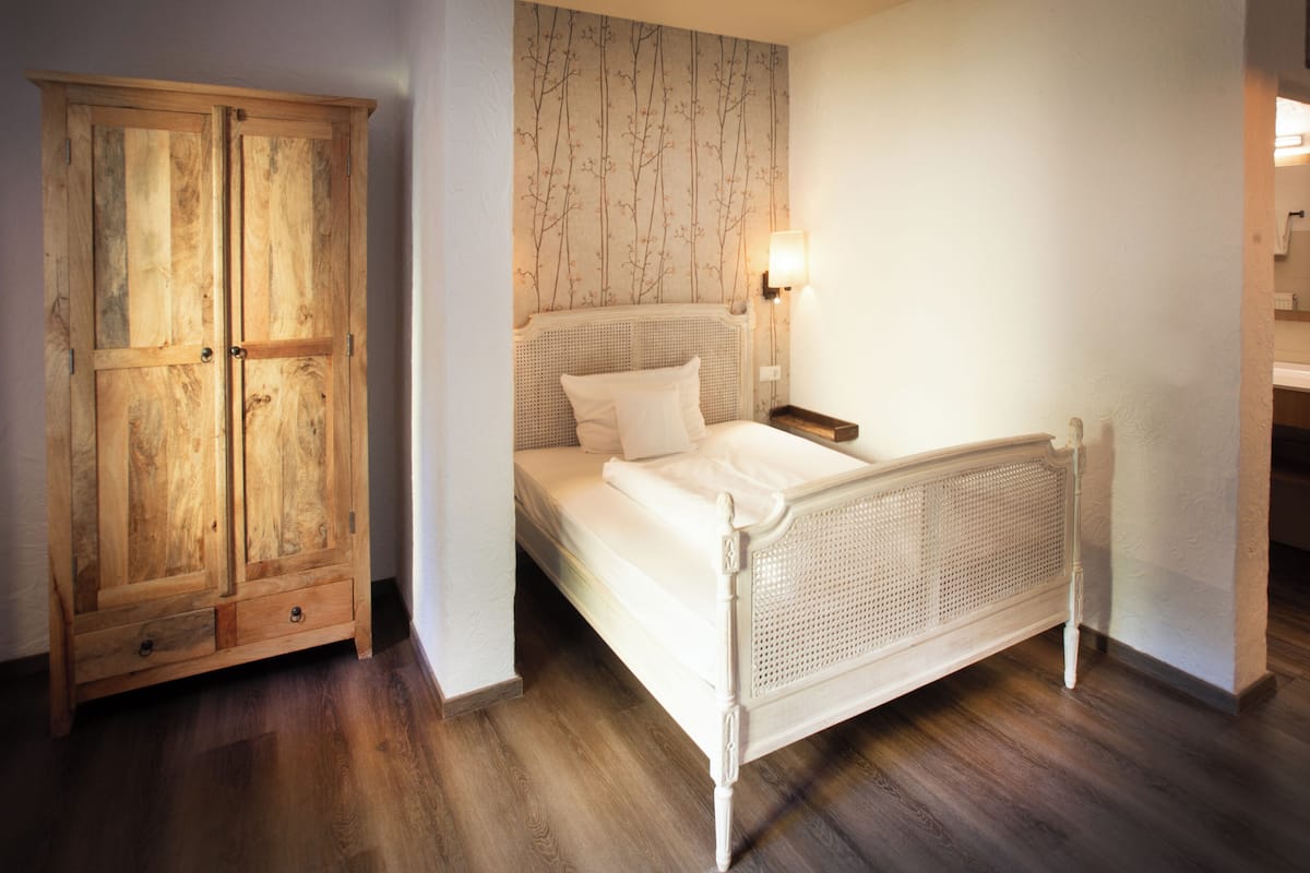 A bedroom with a wooden wardrobe, a white bed with a woven headboard, and a wall-mounted light fixture.