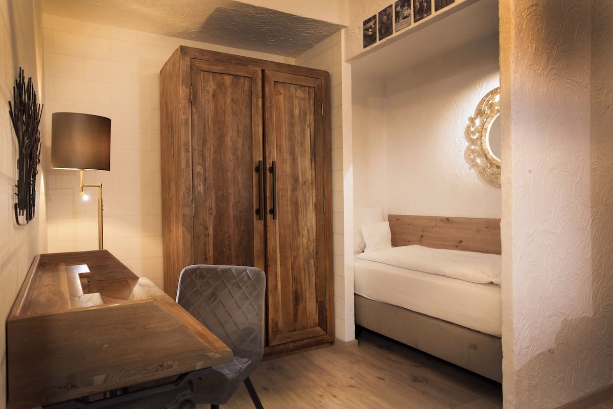 A compact bedroom with a wooden wardrobe, a bed, a desk with a chair, and a wall-mounted lamp.