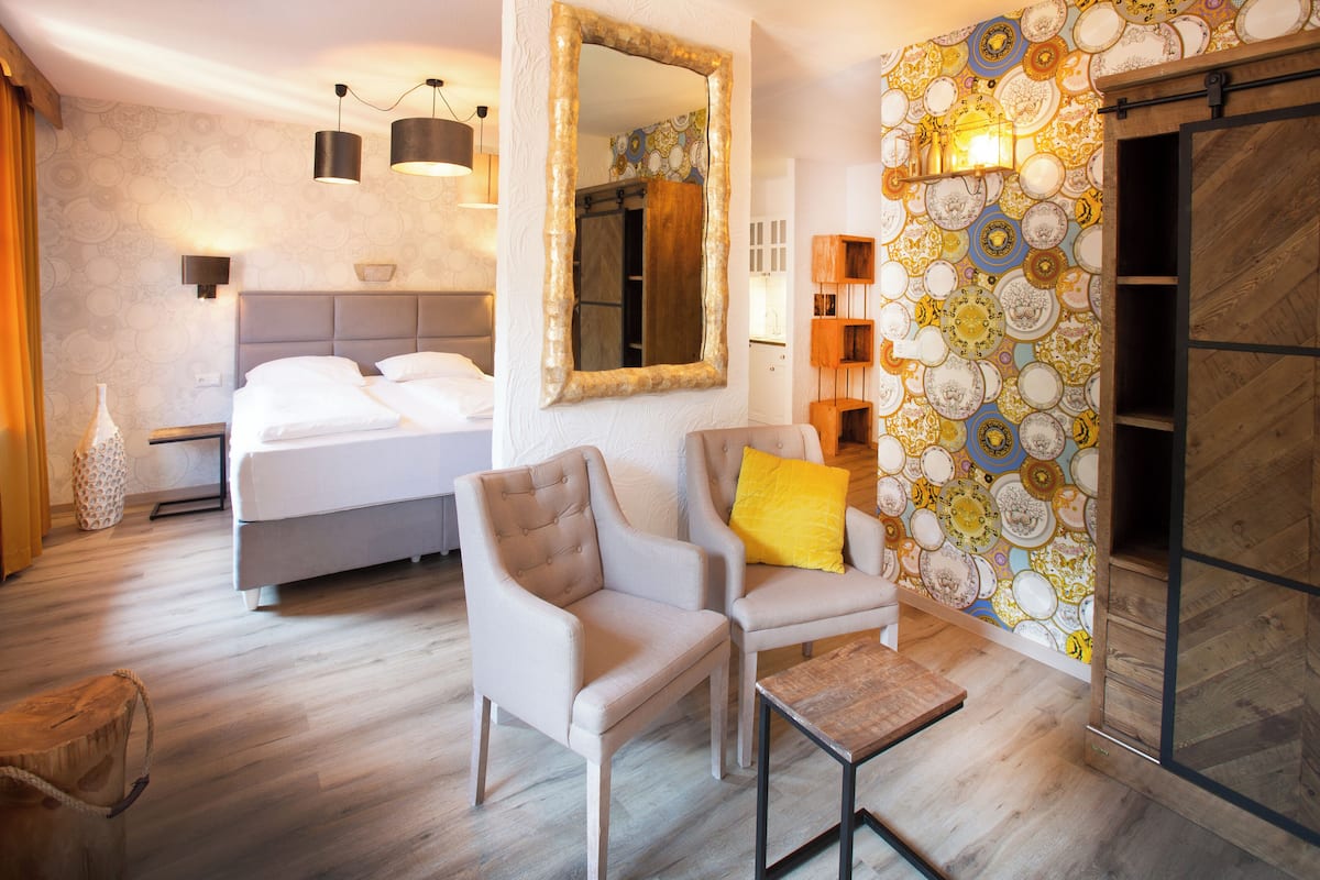 A hotel room with a bed, two armchairs, a small table, a mirror, and a patterned wall.