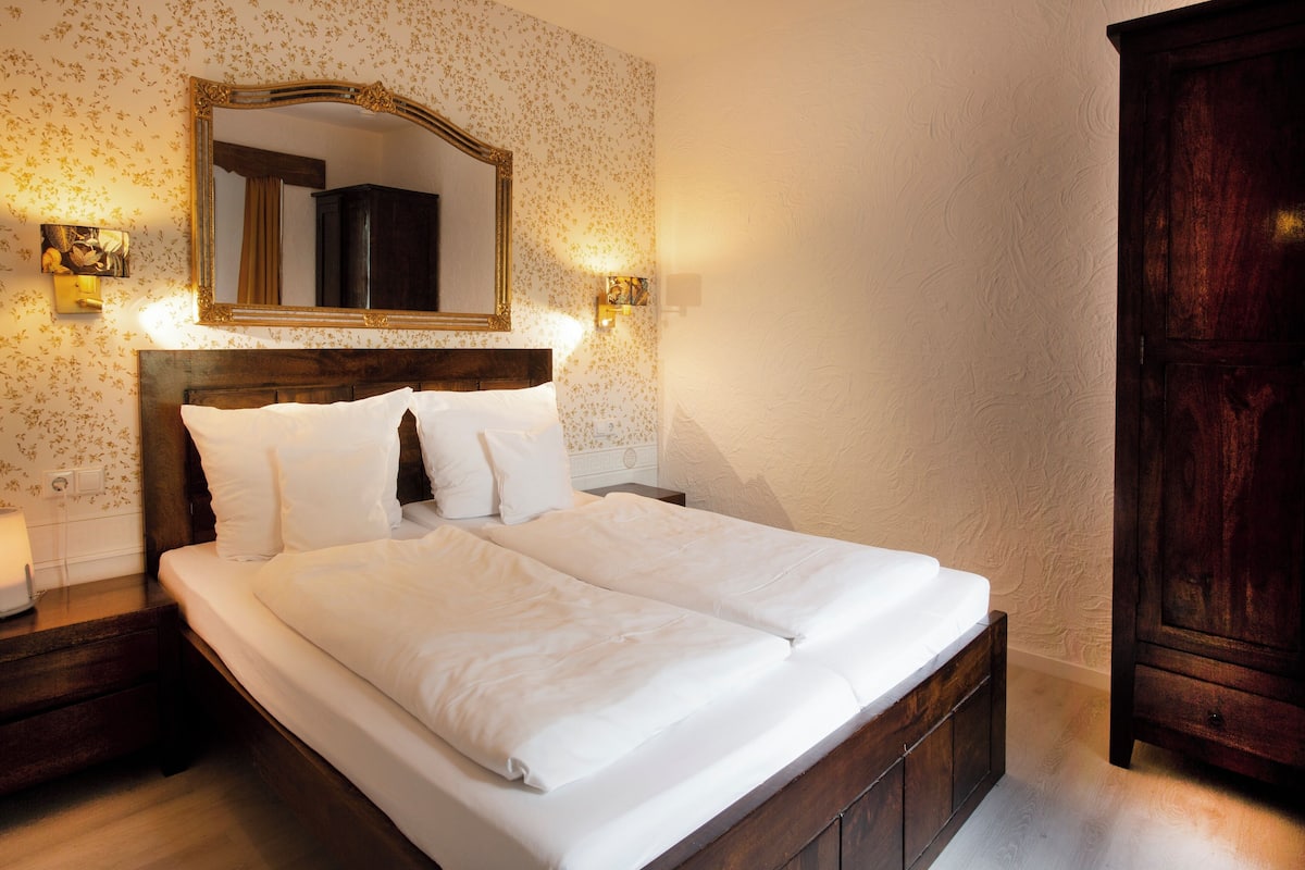 A neatly made bed with white linens, a wooden headboard, and a large mirror above it. There are two wall-mounted light fixtures on either side of the mirror.