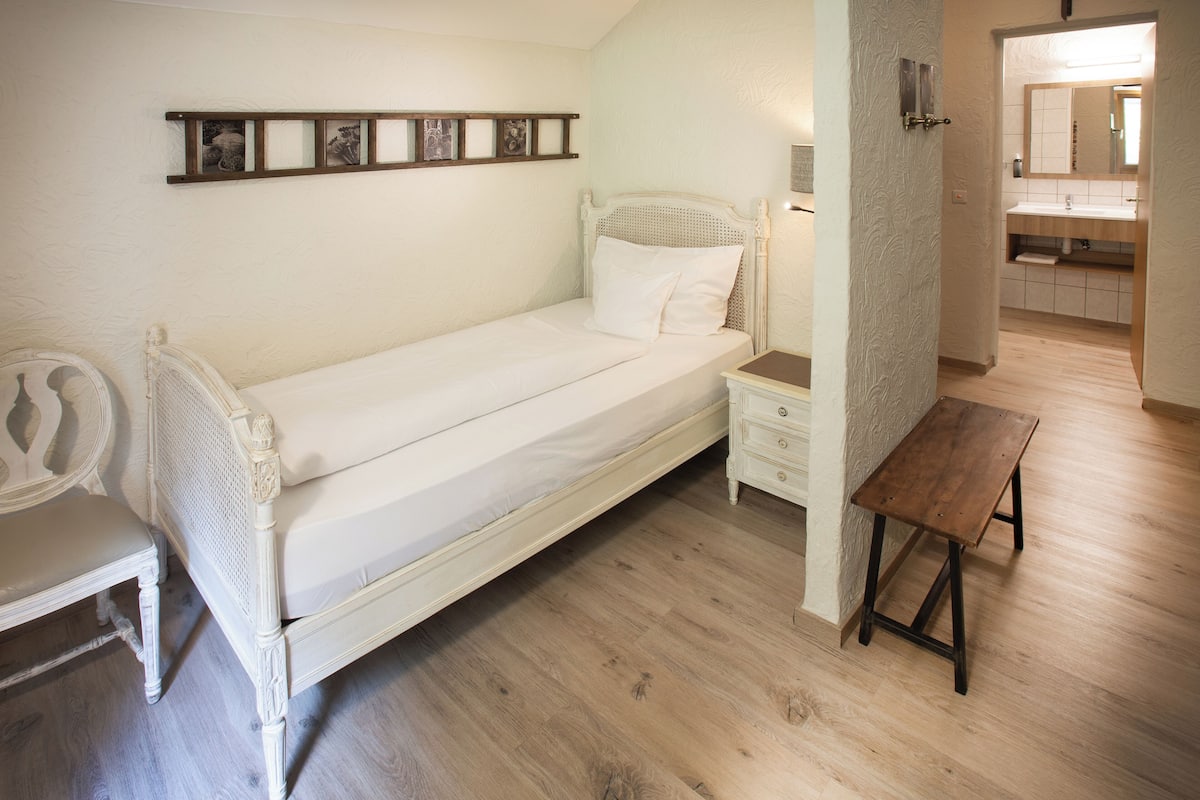 A single bed with a wooden headboard, a bedside table, a bench, and a wall-mounted shelf with pictures.