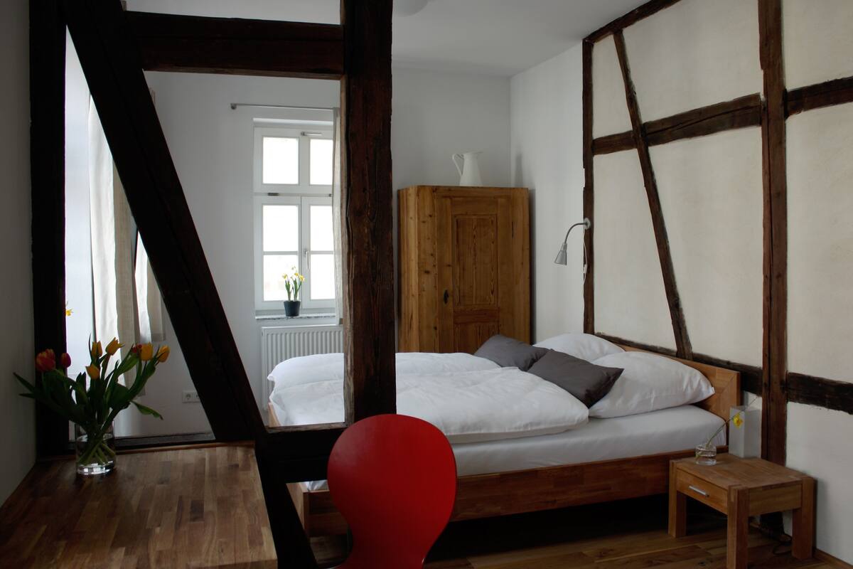 A bedroom with a wooden bed, a red chair, a wooden cabinet, and a window with a view of a white building.