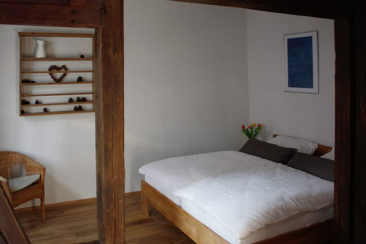 A bedroom with a wooden bed, a wicker chair, a shelf with decorative items, and a framed picture on the wall.