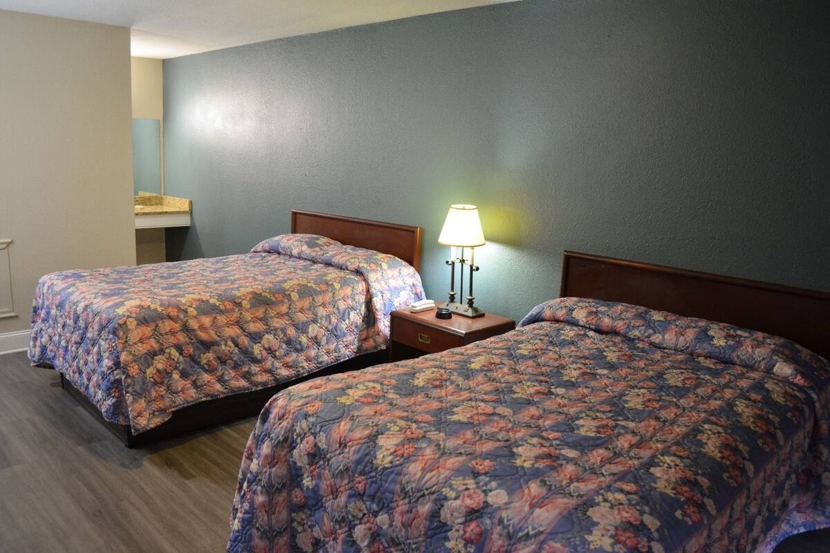 A hotel room with two beds, each with floral bedding, a nightstand with a lamp, and a small table with a mirror.