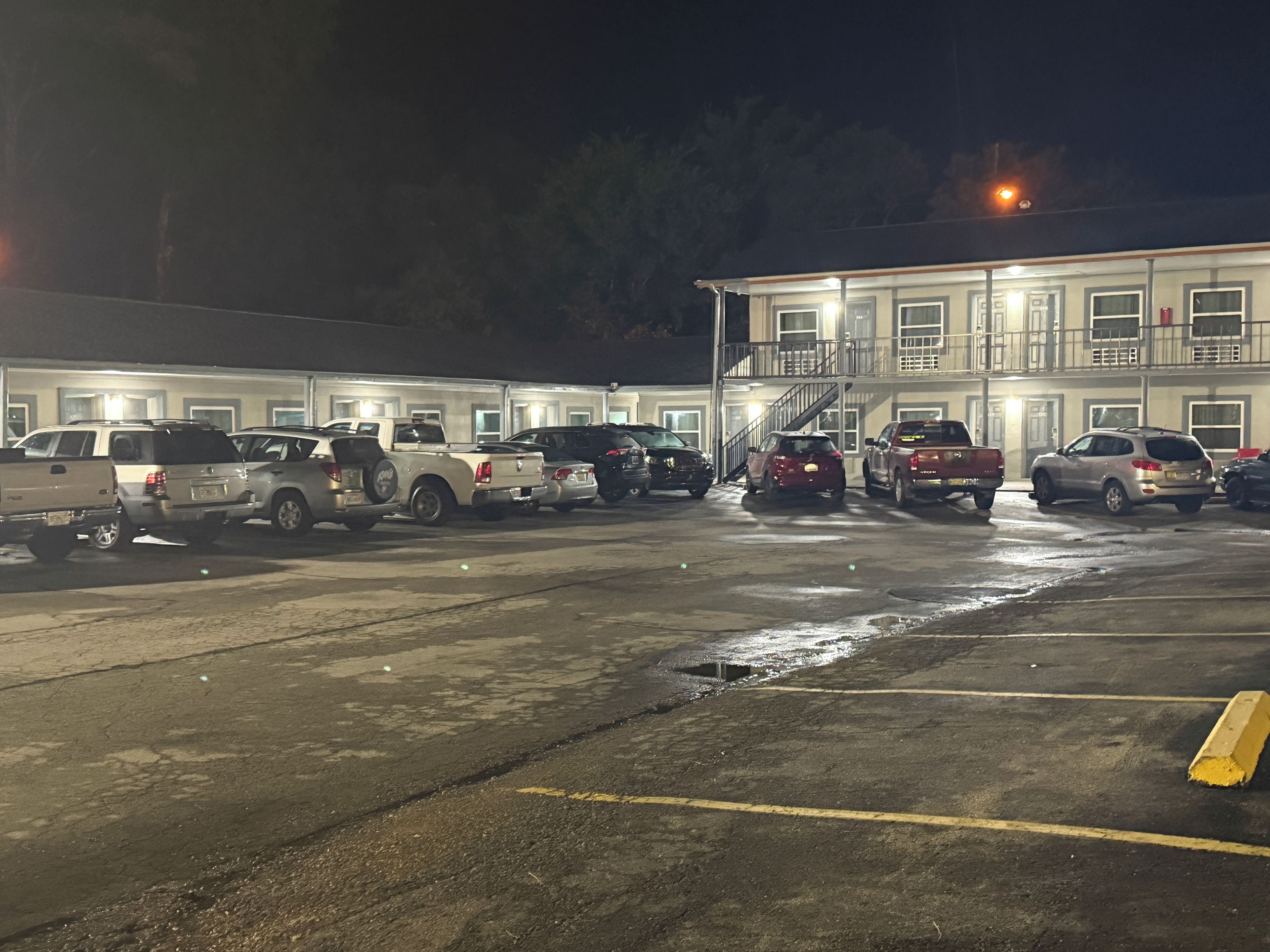Un estacionamiento con varios vehículos, entre ellos camiones y SUVs, frente a un edificio con múltiples ventanales y una escalera que conduce a un nivel superior.