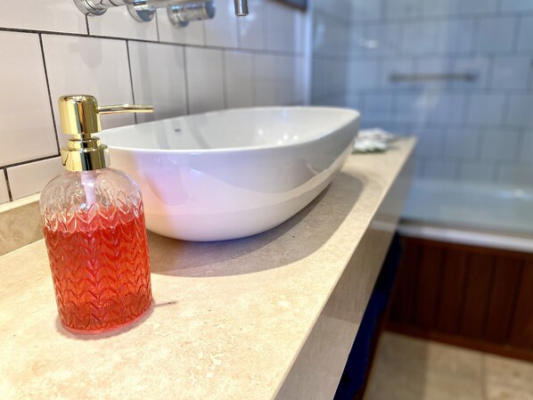 Une salle de bain avec un lavabo blanc, un robinet doré et une bouteille rouge à la texture particulière posée sur le comptoir.