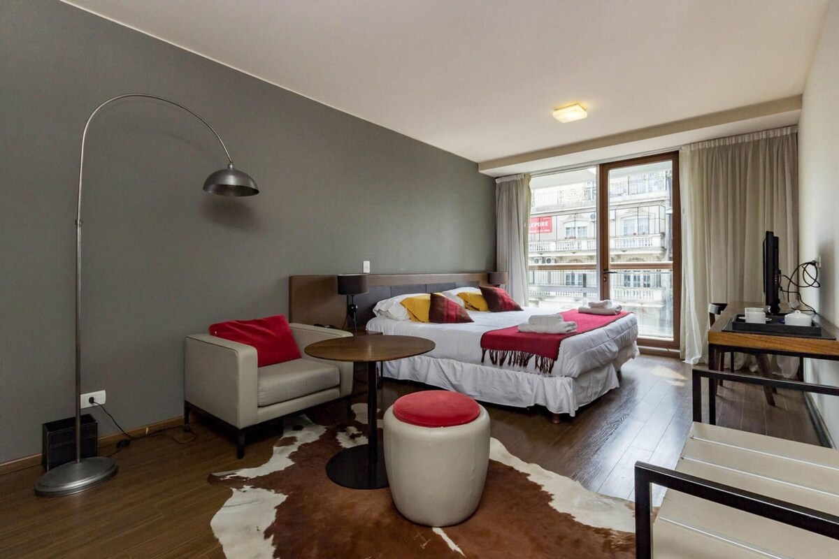 Un hotel moderno con cama, sofá, una mesita, lámpara y vistas a la ciudad a través de amplios ventanales.